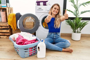 Çamaşır sepetinin yanında oturan genç ve güzel bir kadın elleriyle olumlu bir jest yapmayı onaylıyor, gülümsüyor ve başarı için seviniyor. kazanan jest. 