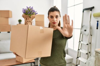 Genç İspanyol kadın, elinde karton bir kutuyla hareket halinde. Ciddi ve kendinden emin bir ifadeyle dur işareti yapıyor. Savunma hareketi. 