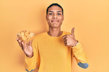 Genç Afro-Amerikalı adam elinde patates cipsi tutarken mutlu ve pozitif gülümsüyor, başparmağıyla mükemmel ve onay işareti yapıyor. 