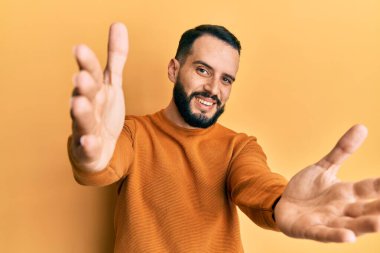 Sakallı, sıradan bir kazak giyen genç bir adam kameraya bakıp sarılmak için kollarını açmış gülümsüyor. Mutluluğu kucaklayan neşeli bir ifade.. 