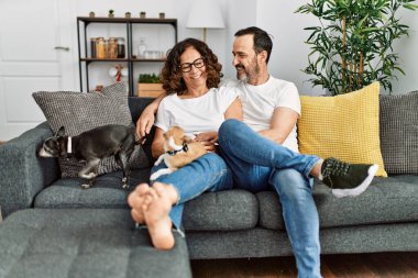 Orta yaşlı, gülümseyen bir çift evde köpeklerle birlikte kanepede oturuyor..