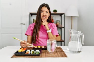 Genç esmer kadın yemek çubuklarıyla suşi yiyor. Bir soru hakkında endişeleniyor ve eli çenesinde endişeli. 