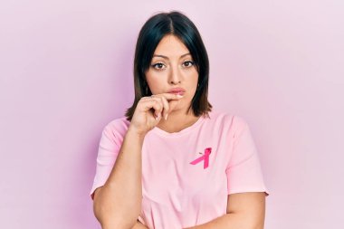 Tişörtüne pembe kanser kurdelesi takan genç İspanyol kadın ağzı ve dudakları parmaklarıyla fermuar gibi kapalı. gizli ve sessiz, tabu konuşması 