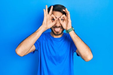 Günlük kıyafetler giyen genç İspanyol adam dürbün gibi jest yapıyor. Dürbün dilini çıkarıyor, gözler parmaklarından bakıyor. çılgın ifade. 