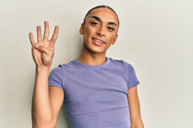 Makyaj yapan, uzun saçlı, günlük tişörtlü İspanyol bir adam dört numarayı gösterip işaret ederken kendinden emin ve mutlu gülümsüyor.. 