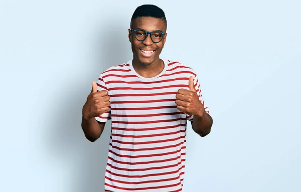 年轻的非洲裔美国人 身穿休闲装 戴着眼镜 成功的标志 手握双手 微笑着竖起大拇指 快乐地做着积极的手势 快乐的表达和胜利的姿态 — 图库照片