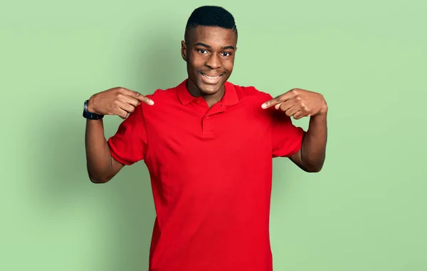 若いアフリカ系アメリカ人の男性は 笑顔で自信を持って見えるカジュアルな赤いTシャツを着て 誇りに思って幸せな指で自分自身を指して — ストック写真