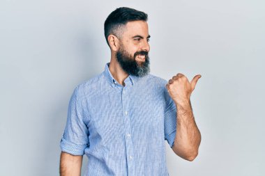 Gündelik kıyafetler giyen, gülümseyen, gülümseyen ve başparmağıyla yan tarafı gösteren İspanyol genç adam.. 