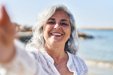 Orta yaşlı bir kadın, kendinden emin gülümsüyor. Sahilde kameranın önünde selfie çekiyor.