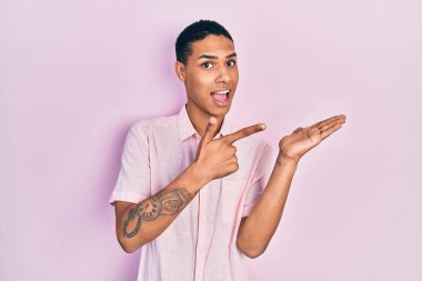 Genç, Afro-Amerikalı, günlük giysiler giymiş. Kameraya hayret ediyor ve gülümsüyor. Elle ve parmakla işaret ediyor.. 