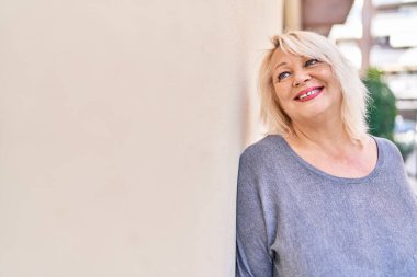 Orta yaşlı sarışın kadın sokakta kendinden emin bir şekilde gülümsüyor.