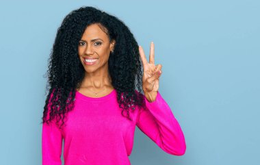 Orta yaşlı Afro-Amerikalı kadın gündelik giysiler giyip iki numarayı gösterirken kendine güvenen ve mutlu bir şekilde gülümsüyor.. 