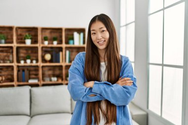 Genç Çinli kız mutlu bir şekilde gülümsüyor kollarını kavuşturup evde duruyor..