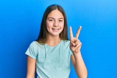 Güzel, esmer, beyaz tişört giyen küçük bir kız. İki numaralı parmaklarıyla onu gösteriyor ve gülümsüyor. Kendinden emin ve mutlu.. 