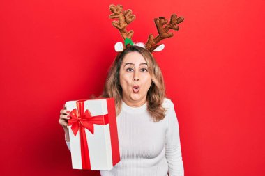 Orta yaşlı, beyaz bir kadın Noel geyiği boynuzları takıyor. Elinde hediyeler var. Şaşırmış ve şaşırmış bir şekilde ağzı açık. 