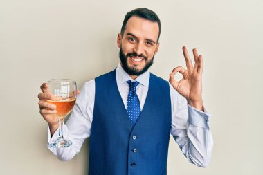 Sakallı genç adam bir bardak beyaz şarap içiyor parmaklarıyla imza atıyor, gülümseyerek mükemmel bir sembol oluşturuyor. 