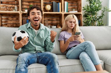 Adam mutlu bir şekilde futbol maçı izliyor ve kız arkadaşı evde akıllı telefon kullanıyor..