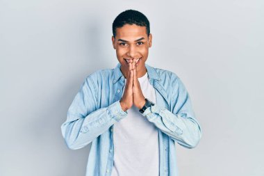 Afro-Amerikan genç adam gündelik giysiler giyip ellerini birleştirip af diliyor kendinden emin bir şekilde gülümseyerek.. 