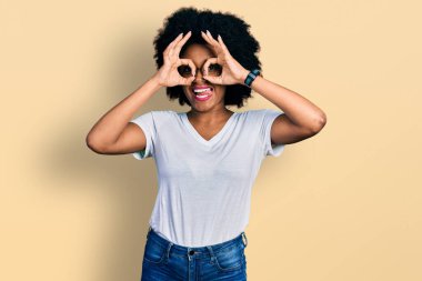 Genç Afro-Amerikalı kadın, günlük beyaz tişört giyiyor dürbün gibi jest yapıyor, dili dışarı çıkarıyor, gözleri parmaklarından bakıyor. çılgın ifade. 