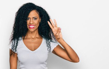 Orta yaşlı Afro-Amerikalı kadın, günlük beyaz tişört giyip üç numarayı gösterirken, kendinden emin ve mutlu bir şekilde gülümsüyor.. 