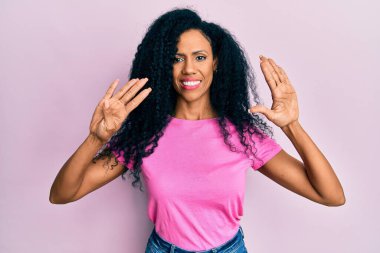 Orta yaşlı Afro-Amerikalı kadın gündelik giysiler giyip dokuz numaralı parmaklarıyla işaret ederken kendinden emin ve mutlu gülümsüyor.. 