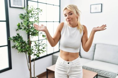 Genç, beyaz bir kadın oturma odasında elleri ve kolları havada, kafası karışmış bir şekilde dikiliyor. şüphe kavramı. 