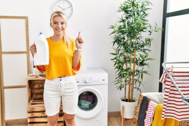 Çamaşır yıkayan, elinde deterjan şişesi olan beyaz bir kadın bir fikir veya soruyla gülümsüyor ve mutlu bir yüzle işaret ediyor. 