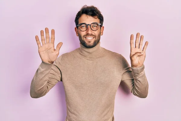 自信と幸せ笑いながら指番号9で表示され 指差す亀甲セーターと眼鏡を身に着けている髭のハンサムな男 — ストック写真
