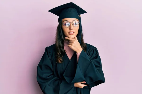 卒業キャップと儀式用のローブを身に着けている若いヒスパニック系の女性は 集中的な表現について考える上で手で 思慮深い顔で笑顔 疑わしい概念 — ストック写真