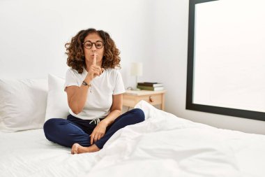 Orta yaşlı İspanyol bir kadın evde yatağında oturuyor ve parmakları dudaklarında sessiz olmak istiyor. sessizlik ve gizli konsept. 