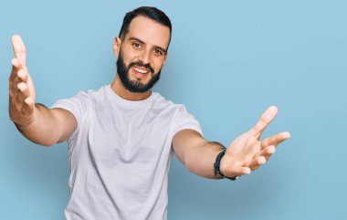 Sakallı, sıradan beyaz tişörtlü genç bir adam kameraya sarılıp gülümsüyor. Mutluluğu kucaklayan neşeli bir ifade.. 