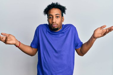 Sakallı, sıradan mavi tişörtlü, kafası karışık, kolları açık, hiçbir fikri olmayan, şüpheli bir yüzü olan Afrikalı genç bir Amerikalı.. 