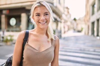 Genç sarışın kız gülümsüyor, mutlu bir şekilde şehirde duruyor.