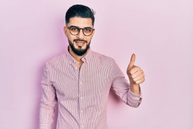 Sakallı, rahat gömlekli ve gözlüklü, mutlu ve pozitif gülümseyen, başparmağıyla mükemmel ve onay işareti yapan yakışıklı bir adam. 