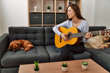 Genç İspanyol kadın klasik gitar çalıyor. Evde köpeklerle birlikte kanepede oturuyor.