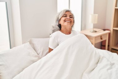 Orta yaşlı kadın yatakta kendinden emin bir şekilde gülümsüyor.