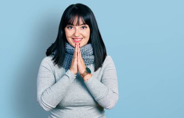 Latin kökenli iri yarı, kış eşarbı takan, elleriyle dua eden, affedilmek isteyen, kendinden emin gülümseyen bir kadın.. 