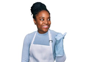 Önlük giymiş, gülümseyen, gülümseyen ve başparmağıyla yüzünü gösteren Afro-Amerikan genç bir kadın.. 