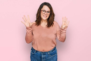 Genç ve cüsseli kadın günlük kıyafetler giyiyor ve gözlük takıyor dokuz numaralı parmaklarıyla işaret ediyor ve gülümsüyor kendinden emin ve mutlu.. 