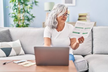 Orta yaşlı muhasebe kadını evdeki koltukta dizüstü bilgisayar kullanıyor.