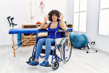 Fizyoterapi kliniğinde tekerlekli sandalyede oturan genç orta doğulu kadın dokuz numarayı gösterip işaret ederken kendinden emin ve mutlu bir şekilde gülümsüyor.. 