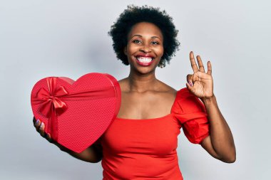 Sevgililer günü hediyesini tutan Afrikalı Amerikalı genç bir kadın parmaklarıyla imza atıyor, gülümseyerek mükemmel bir sembol oluşturuyor. 