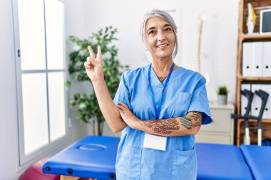 Tıp kliniğinde fizyoterapist üniforması giyen, kameraya gülümseyen, zafer işareti yapan orta yaşlı, gri saçlı bir kadın. İki numara.. 