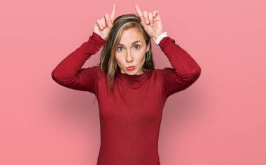 Genç, sarışın bir kadın, günlük kıyafetler giyiyor, komik jestler yapıyor, parmakları kafasının üstünde boğa boynuzu gibi. 