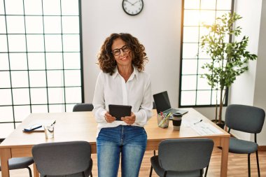 Orta yaşlı İspanyol kadın, ofiste dokunmatik ped kullanarak çalışırken kendine güvenerek gülümsüyor.