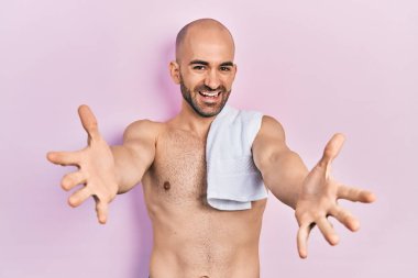 Genç kel adam üstsüz olarak kameraya bakıyor ve sarılmak için kollarını açmış gülümsüyor. Mutluluğu kucaklayan neşeli bir ifade.. 