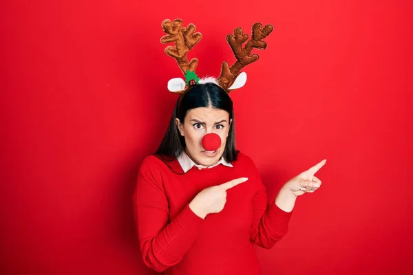 若いですヒスパニック系の女性を身に着けています鹿のクリスマスの帽子と赤い鼻心配脇と両手で神経を指しています 懸念と驚きの表情 — ストック写真