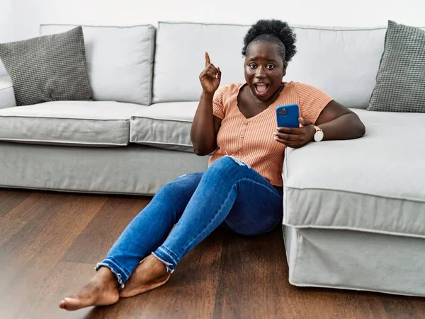Young African Woman Using Smartphone Sitting Floor Home Pointing Finger — стоковое фото