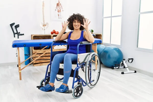 Fizyoterapi kliniğinde tekerlekli sandalyede oturan genç orta doğulu kadın dokuz numarayı gösterip işaret ederken kendinden emin ve mutlu bir şekilde gülümsüyor.. 