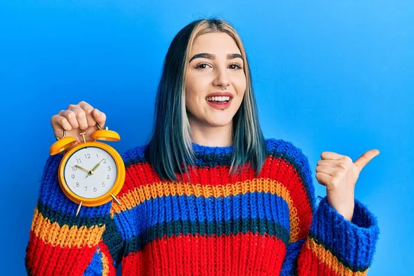 Jovem Menina Moderna Segurando Despertador Apontando Polegar Até Lado Sorrindo — Fotografia de Stock